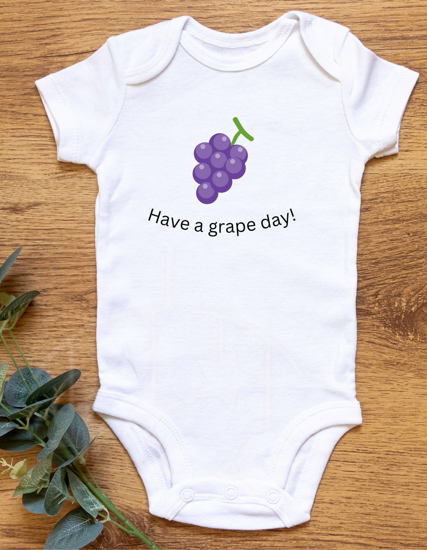 White onesie that says have a grape day and has an image of a bunch of grapes on a wood background with green leaves.