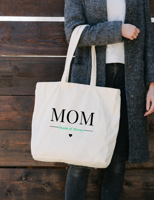 White lady with wearing jeans and a gray jacket with a square canvas tote with MOM in capital letters with Made of Money underneath with a heart hanging from her arm.