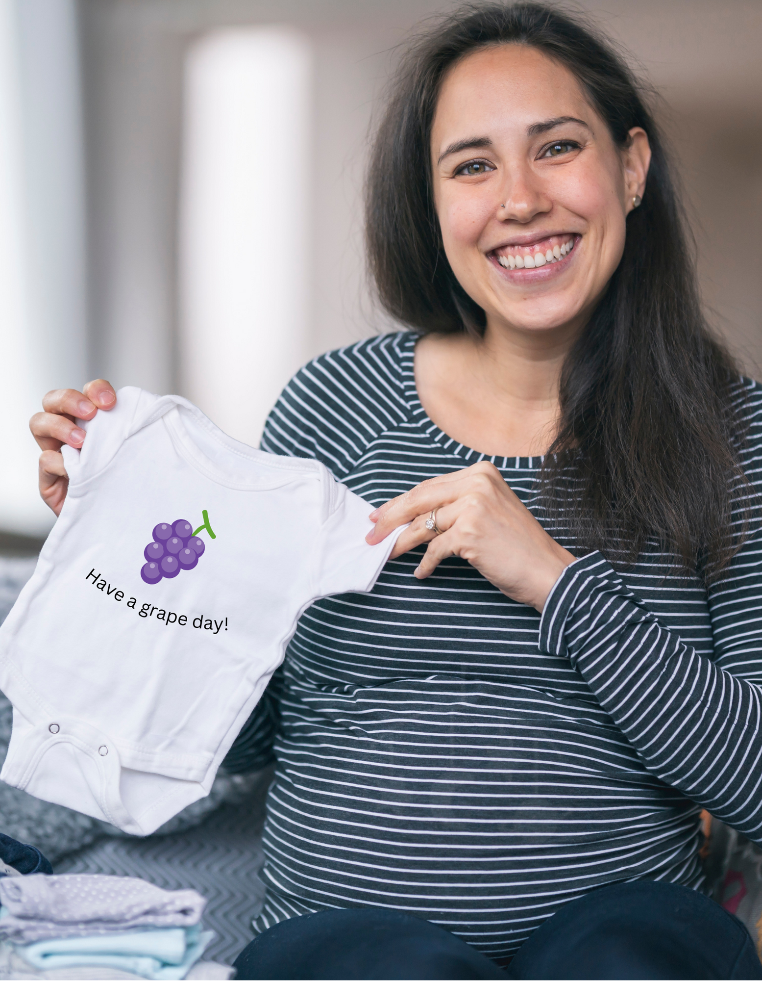 A pregnant women with brown hair, smiling, holding a white baby onesie that says have a grape day and has an image of a bunch of grapes.