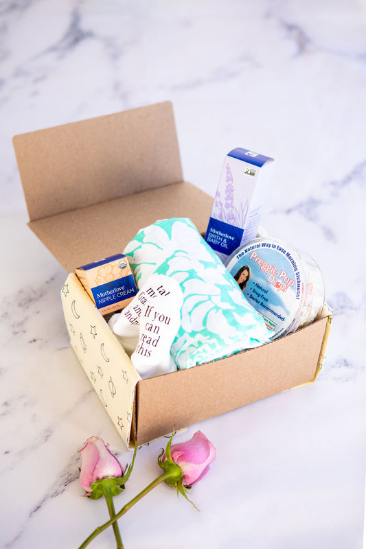 picture of the expectant mom box that has white baby socks that read if you can read this take me to grandma and grandpas, preggie pop drops, a teal and white swaddle, and motherlove nipple cream and birth and baby oil resting on a marble background with two pink roses in the left hand side