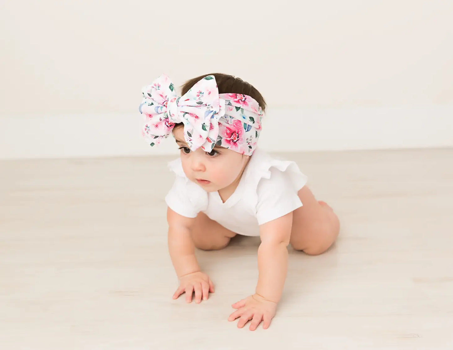 Picture of a white baby with brown hair in a white ruffle onesie crawling wearing a big bow headband that is white with pink flowers and has blue and green leaves