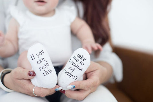 a picture of a white baby in a white top where white socks that say if you can read this take me to grandma and grandpa's on the sole of the sock and the baby is sitting on a womens lap 