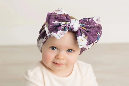 Picture of a white baby girl in a white shirt wearing a big bow headband that has a purple background with white flowers and other colors like yellow, black, blue