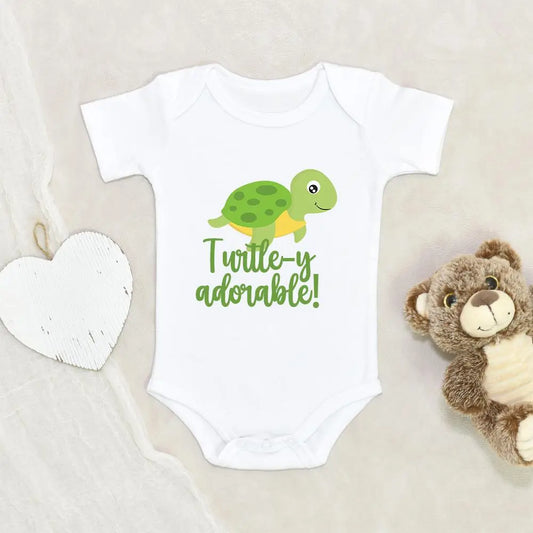 image of a white baby onesie that has an image of a green turtle with a smiling face that has the saying turtle-y adorable! on a marble background with a white wood heart and a brown teddy bear.