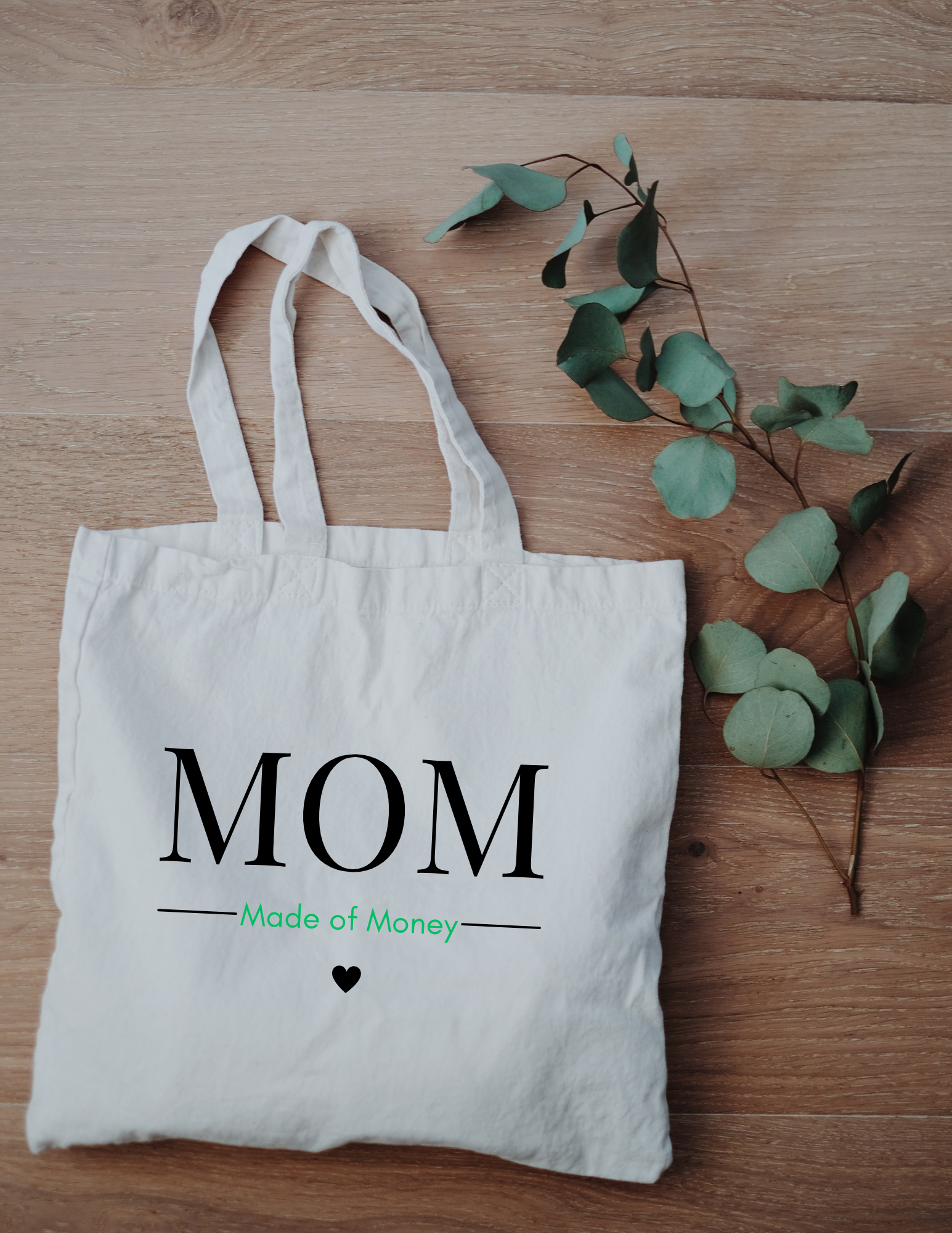 Square canvas tote with MOM in capital letters with Made of Money underneath with a heart below that on a wood background with a eucalyptus leaf.