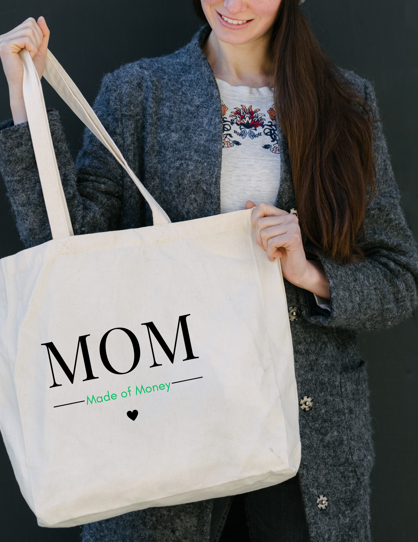 White lady with long brown hair in jeans and a gray jacket holding a square canvas tote with MOM in capital letters with Made of Money underneath with a heart below that.