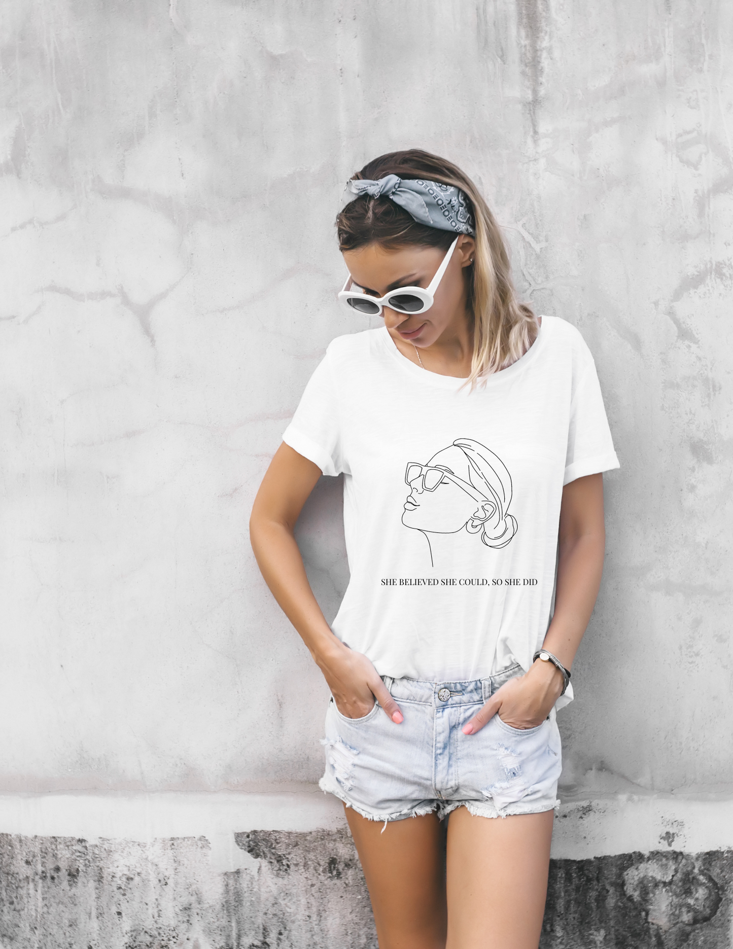 A white women with blond hair, wearing  a white t-shirt with a black outline of a women wearing sunglasses with her hair in a bun and earrings that has a quote she believed she could, so she did in a pair of jean shorts with sunglasses and headband.
