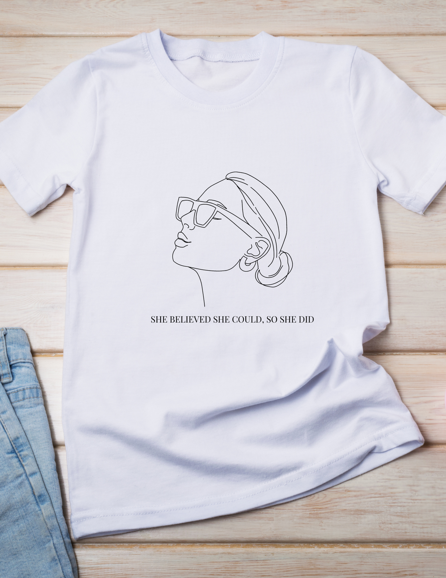 Women's white t-shirt with a black outline of a women wearing sunglasses with her hair in a bun and earrings that has a quote she believed she could, so she did underneath on a wood background with an image of some jeans.