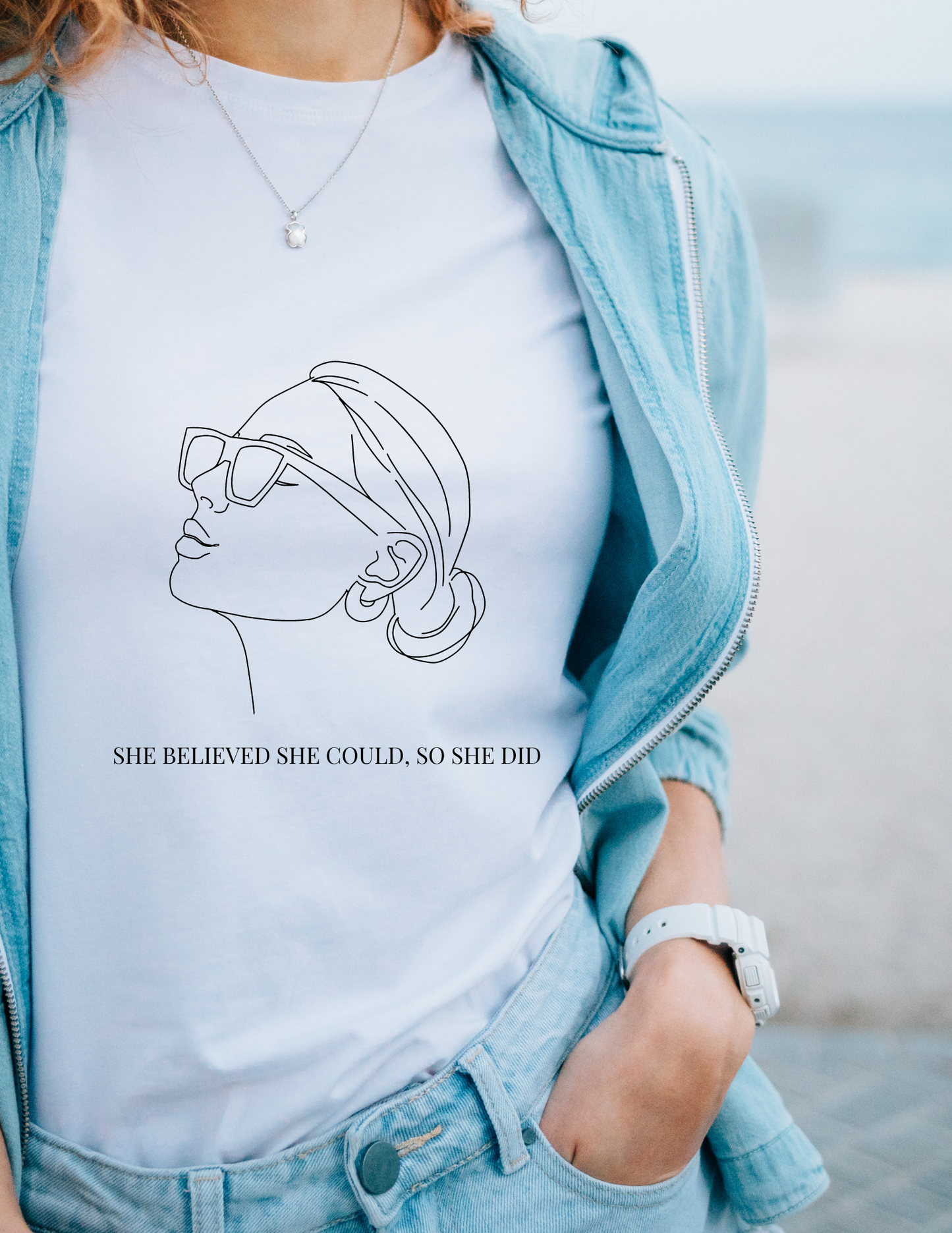 A women's upper body wearing a white t-shirt with a black outline of a women wearing sunglasses with her hair in a bun and earrings that has a quote she believed she could, so she did with a blue jacket and jeans.