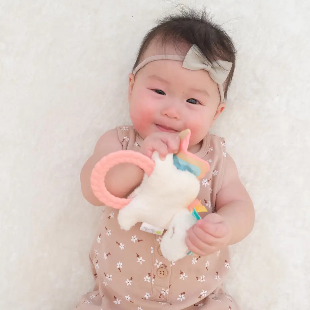 picture of a asian baby girl wearing a tan bow headband and a tan flower dress holding the unicorn rattle on a white blanket background